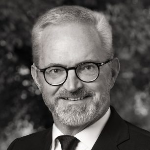 En man med glasögon och skägg ler, klädd i kostym och slips. Bakgrunden är suddig och skapar ett professionellt intryck.