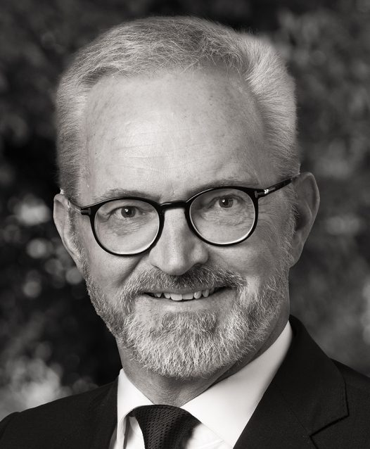 En man med glasögon och skägg ler, klädd i kostym och slips. Bakgrunden är suddig och skapar ett professionellt intryck.